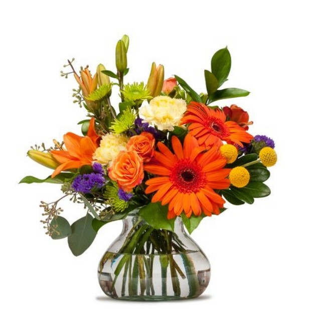 Bright floral arrangement featuring orange gerbera, lilies, roses, and purple and yellow accent blooms in a glass vase.