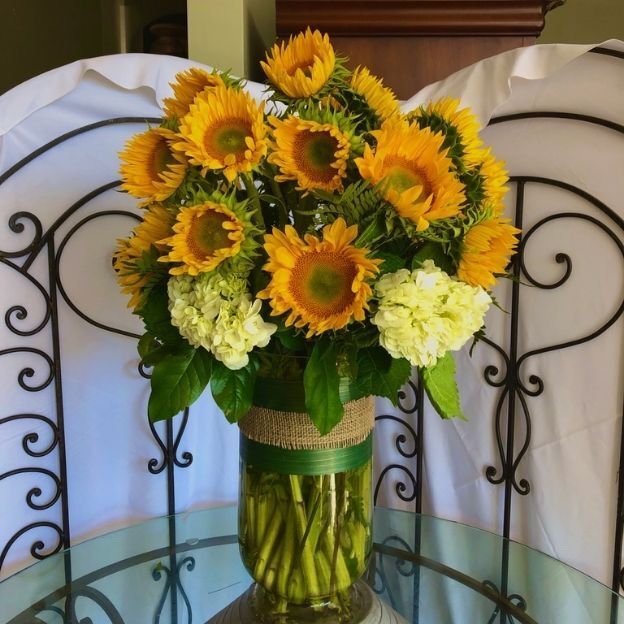 Bright sunflower arrangement with white hydrangeas and green foliage in a clear vase with burlap ribbon.